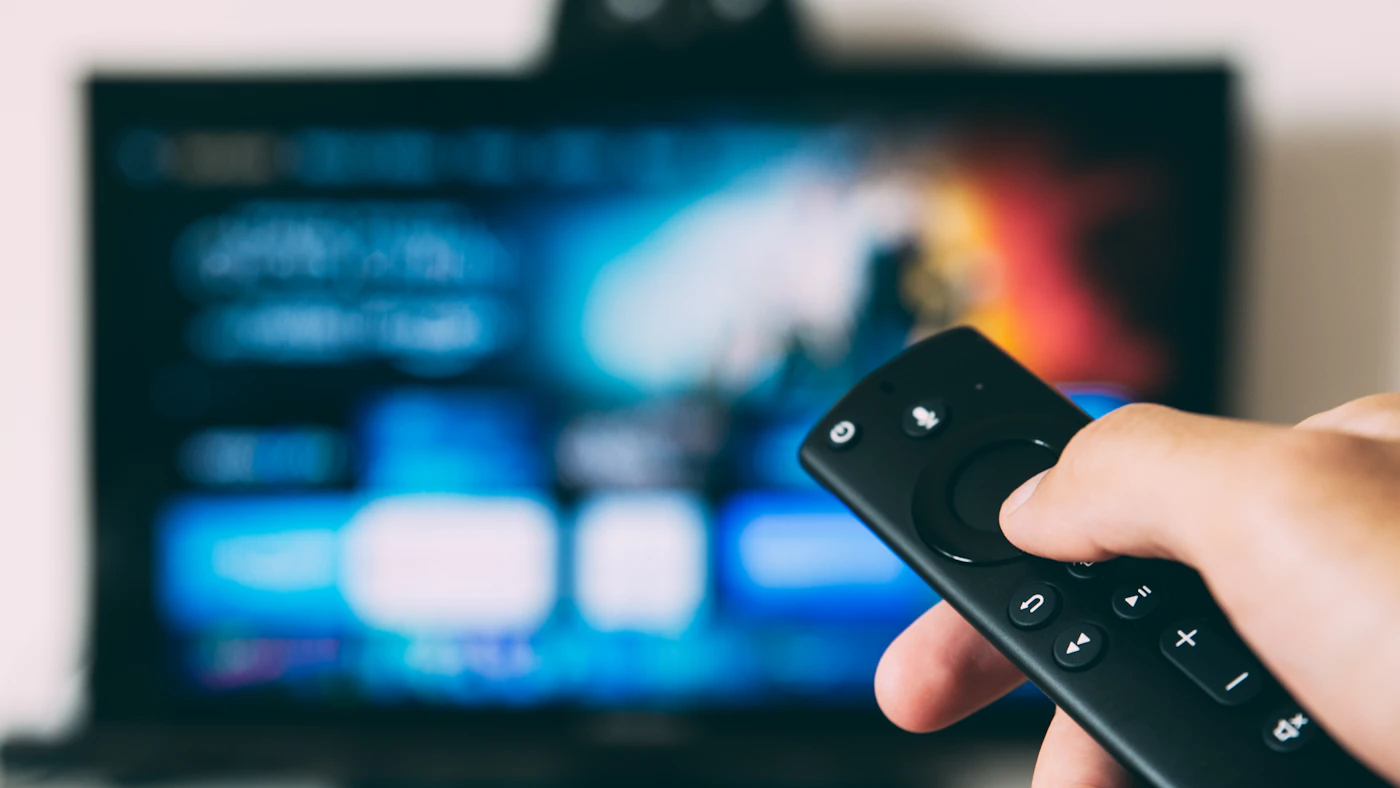 Hand holding a streaming remote in front of a blurred TV screen